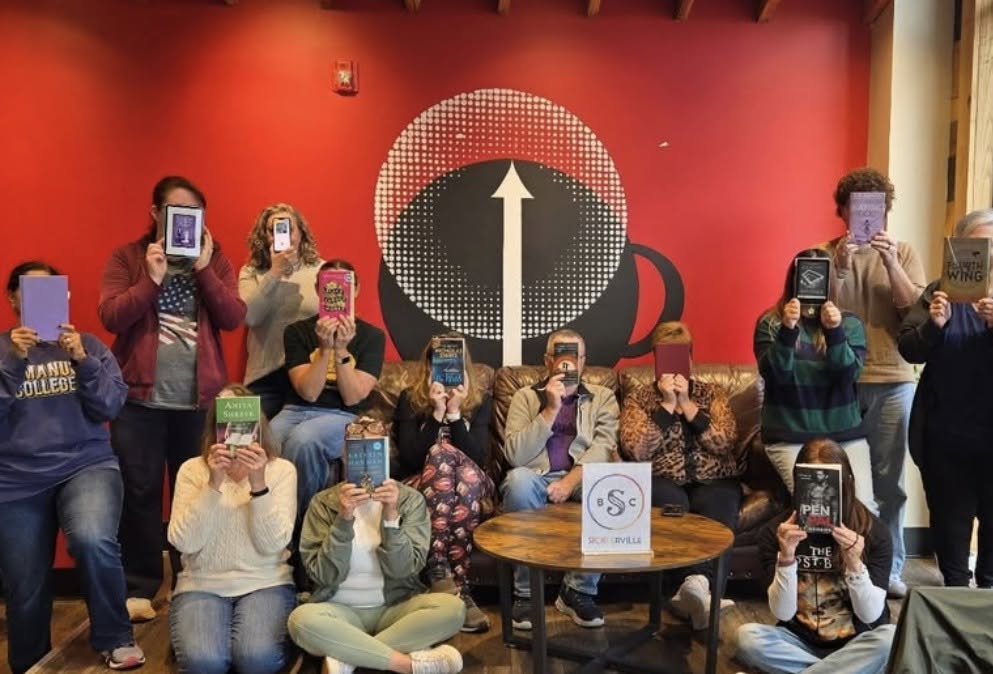 Book club members holding up their books in front of the High Grounds cup mural