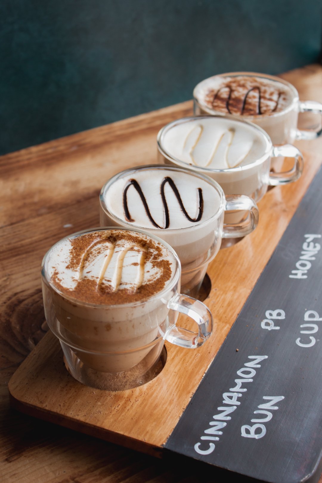 Coffee flight of lattes labeled Cinnamon Bun, PB Cup and Honey on a wooden board
