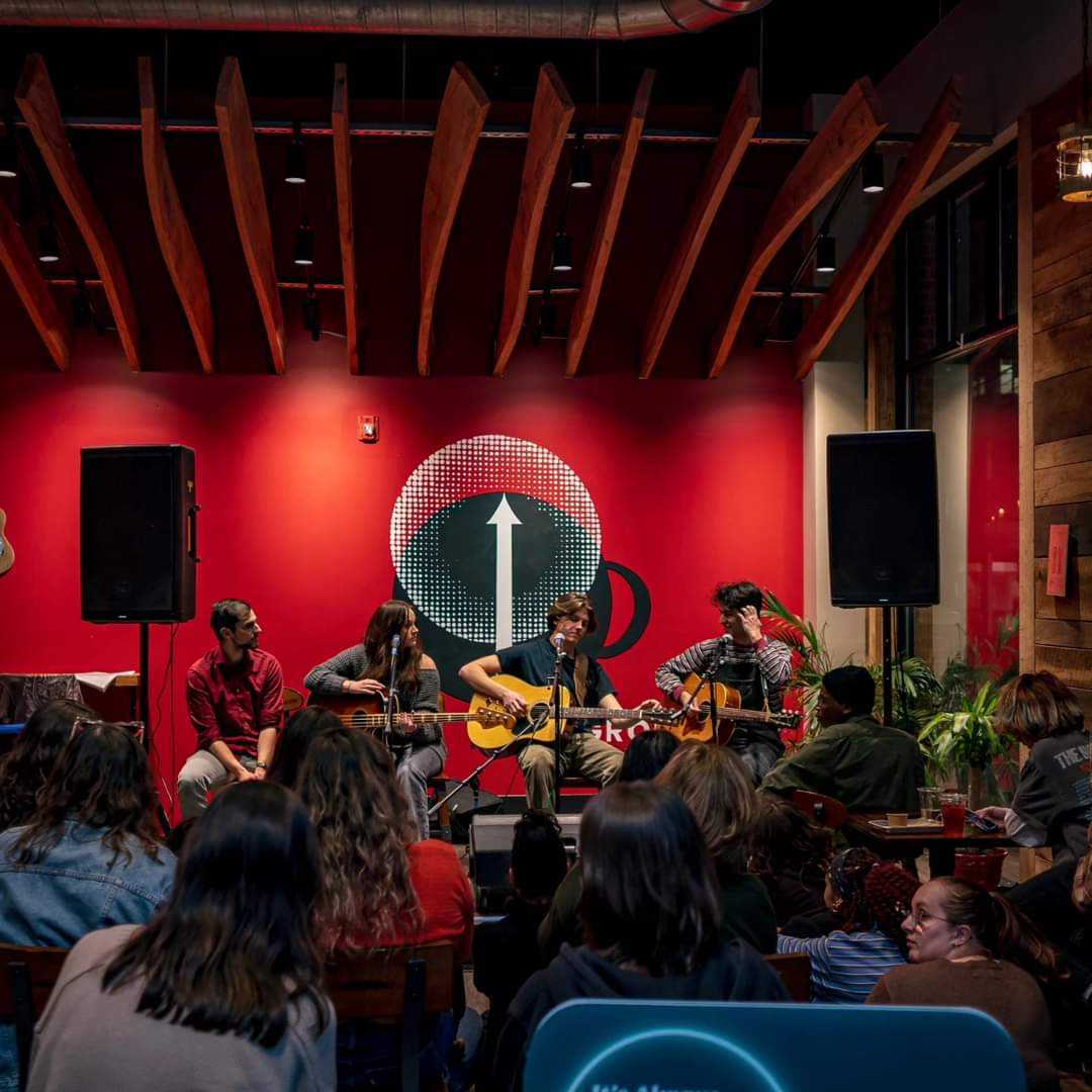 Acoustic musicians performing in front of the red High Grounds mural to a packed room